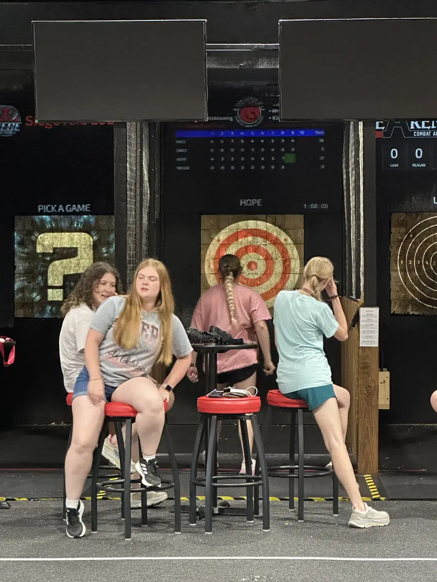 Four young people on red stools at an axe lane, with a bullseye target on the boards