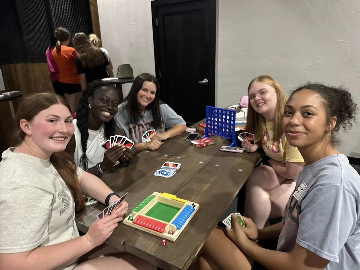 Five young people playing cards and board games at a wooden table, smiling at the camera